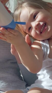 A happy toddler boy with blonde hair and a big smile applies lotion to her hands with the help of an adult. Bright natural lighting, a cozy home setting, and a joyful moment of self-care