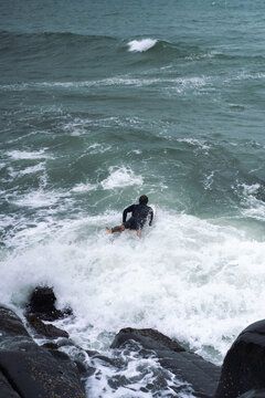 Man in a wetsuit jumps from rocks into the ocean with his surfboard