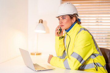 Young woman builder in safety jacket working in office while talking on phone and using laptop showcasing professionalism in construction environment