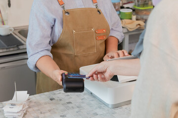 Senior entrepreneur cafe owner barista holds POS terminal while customer swipes creditcard, Asian retired female in apron smiling at counter, demonstrating cashless payment inside modern coffee shop