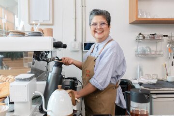 Senior entrepreneur cafe owner barista chats with female customer at counter, Asian male in apron smiling while presenting croissant plate, espresso machine and shelving frame cozy evening scene