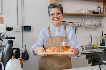 Senior entrepreneur cafe owner barista presents bakery croissants on tray, Asian retired female in apron grins across counter, grinder and shelves create welcoming small business hospitality scene.