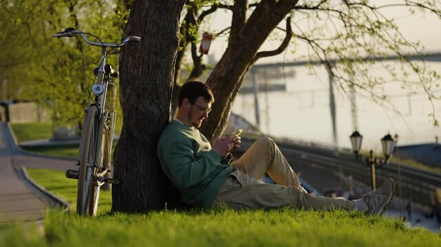Young man leisurely browsing dating app on smartphone while sitting under tree, bicycle parked nearby river in peaceful park setting during warm summer day
