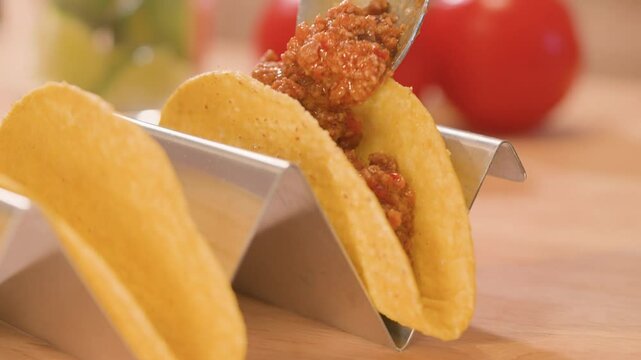 Close-up of hard corn taco shells, partially filled with seasoned meat and a spoonful being added, on a stainless steel taco stand with one empty shell.