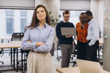 Confident businesswoman posing with crossed arms in modern office with colleagues working
