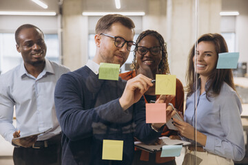 Diverse group of professionals brainstorming with sticky notes on a glass wall, fostering creativity and teamwork in a modern office setting