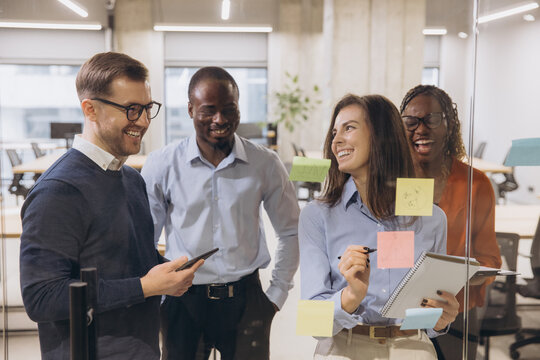Diverse group of professionals brainstorming with sticky notes on a glass wall, fostering creativity and teamwork in a contemporary office environment