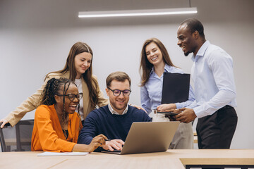 Diverse professional team collaborating, reviewing laptop data with shared enthusiasm in modern office environment, displaying teamwork spirit