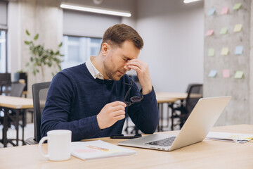 Tired office male worker suffering from eye strain, taking off glasses and rubbing his eyes after...
