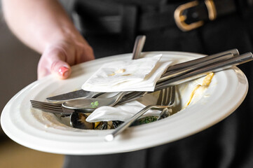 A close-up of a hand holding a dirty plate with used cutlery and napkins, representing table clearing after a meal. Food scraps and mussel shells remain on the dish, illustrating restaurant service