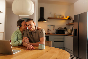 Young multi ethnic couple in love are embracing and holding hands, sitting at wooden kitchen table with laptop and cup of coffee or tea, enjoying time together at home
