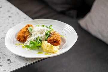 A gourmet dish of breaded fried meat with mashed potatoes, fresh green salad, and grated cheese, garnished with green sauce on a white plate, presented on a speckled countertop.