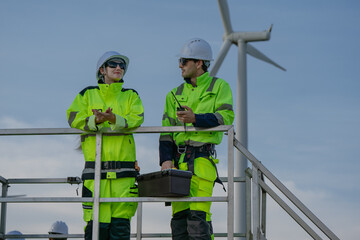 Maintenance engineer team standing at windmills at wind turbine farm. Group of people wear safety helmet and uniform working at alternative renewable energy wind station. Sustainable energy technology