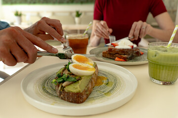 Hands cutting avocado toast with boiled eggs, healthy breakfast or brunch in a cafe with matcha latte