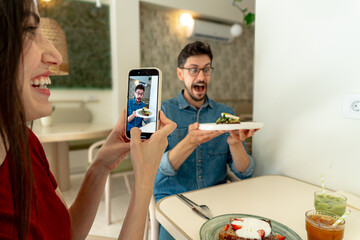 Woman taking pictures with smartphone of man making funny face and holding plate with toast in cafe