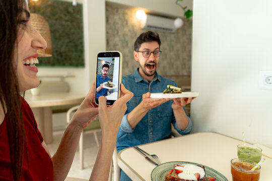 Woman taking pictures with smartphone of man making funny face and holding plate with toast in cafe