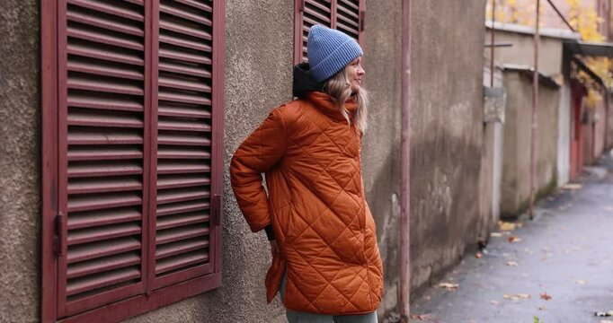 Young woman in orange quilted coat and blue beanie searching through pockets of coat. Moment of minor inconvenience on damp city street slow motion