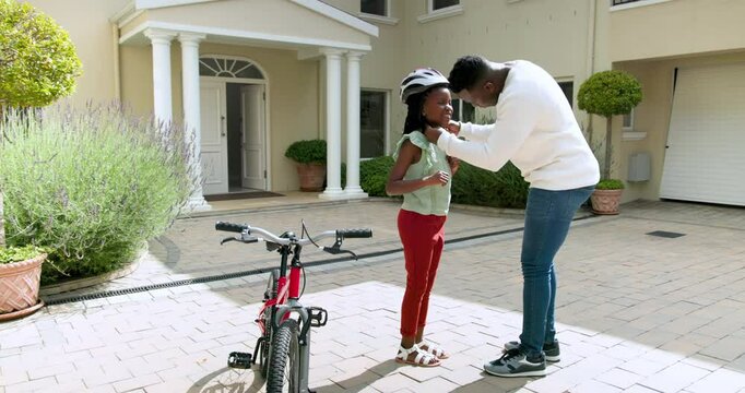 Happy black father teaching his smiling daughter to ride a bike outside their home, girl wearing hel