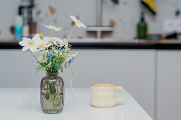 A floral still life with flowers in a vase and a mug on a white table.