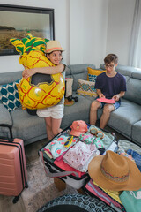 Happy girl having fun hugging inflatable pineapple toy and smiling while brother playing with ping pong paddles. Siblings preparing suitcase for summer vacation, radiating joy and excitement for trip.