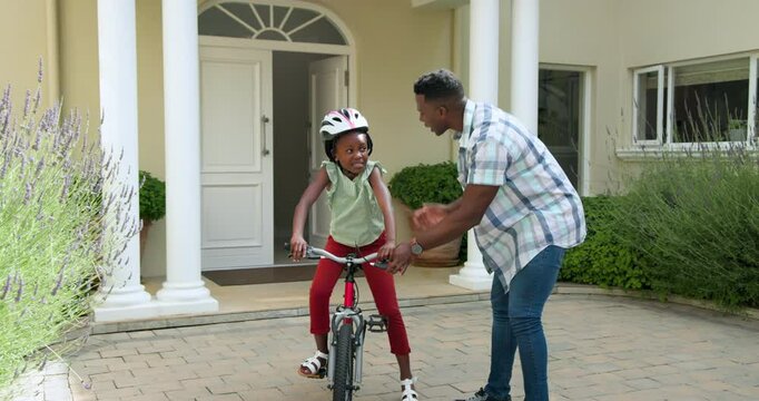 Happy black father teaching his smiling daughter to ride a bike outside their home, girl wearing hel