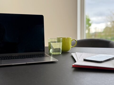 Close-up of an open laptop computer with a blank screen, a mobile phone, paperwork and drinks on a dining table