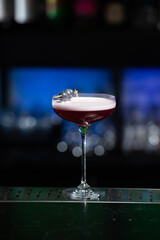 purple cocktail with a foamy white top is garnished with skewered blueberries on a textured bar surface