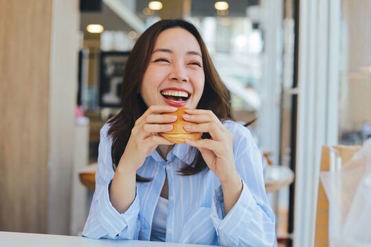 A cheerful young Asian woman is smiling brightly while holding a burger with both hands, ready to take a bite. She is enjoying her meal at a modern cafe or restaurant.