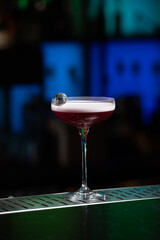 purple cocktail with a foamy white top is garnished with skewered blueberries on a textured bar surface