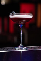 purple cocktail with a foamy white top is garnished with skewered blueberries on a textured bar surface