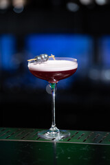 purple cocktail with a foamy white top is garnished with skewered blueberries on a textured bar surface
