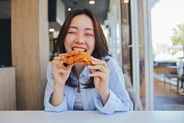 A joyful young Asian woman enjoying a piece of fried chicken at a modern restaurant. She laughs...