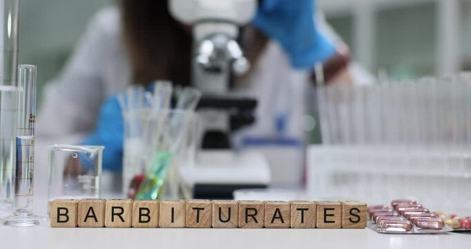 Text Barbiturates of wooden cubes against assistant in laboratory. Woman scientist works with microscope surrounded by glassware and blisters in lab
