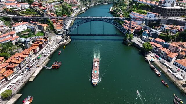 Aerial of a river cruise ship sailing beneath the iconic Dom Lu&iacute;s I Bridge in Porto, surrounded by historic buildings and bustling riverbanks
