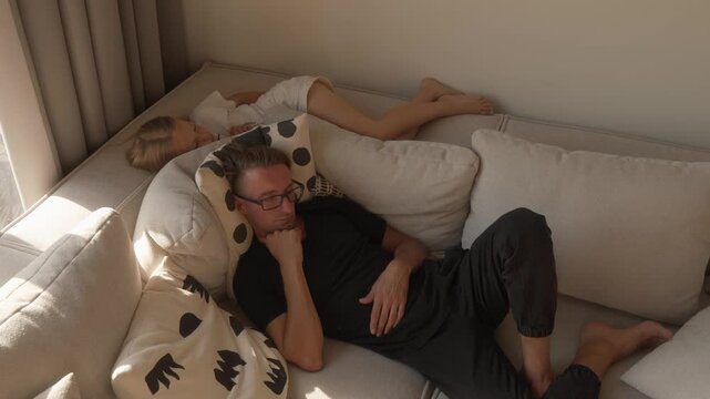 A father and his young daughter sleep peacefully side-by-side on a comfortable sofa in their living room. This serene scene captures a moment of shared rest, family warmth, and tranquility at home.