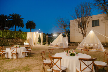 Outdoor wedding reception setup with draped tents, round tables, and ambient lighting creates an...