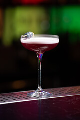 purple cocktail with a foamy white top is garnished with skewered blueberries on a textured bar surface