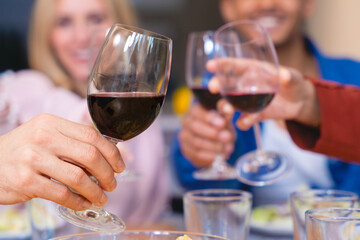 Close-up of unrecognizable happy friends toasting with red wine at a cheerful dinner party. Celebration, friendship, and good food in a warm and vibrant atmosphere at the table. 