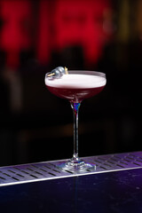 purple cocktail with a foamy white top is garnished with skewered blueberries on a textured bar surface