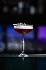 purple cocktail with a foamy white top is garnished with skewered blueberries on a textured bar surface