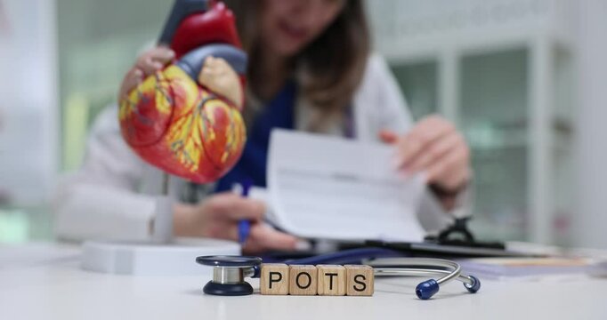 Abbreviation POTS on wood blocks with heart model against working doctor. Cardiologist analyzes papers about postural orthostatic tachycardia syndrome