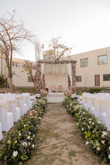 Elegant outdoor wedding setup with floral aisle, decorated stage, and rows of white chairs on a grassy lawn. A dreamy and romantic ambiance.