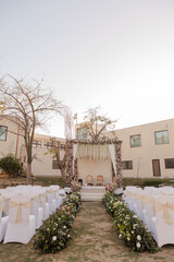 Outdoor wedding setup featuring a floral mandap, white chairs, and a flower-lined aisle, set against a building backdrop. Ceremony in a garden setting.