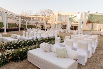 Outdoor event venue setup with white chairs, sofas, floral arrangements, and a draped canopy against a modern building in natural light.