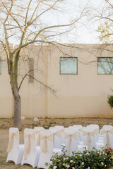 Outdoor wedding setting with elegant white chairs adorned with decorative ties, set against a backdrop of a modern building and a bare tree.