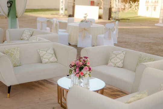 Outdoor event lounge area with white couches, decorative pillows, and floral centerpiece on a round table, set against a backdrop of white event furniture.
