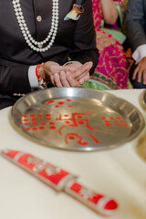 A ritual at a wedding ceremony. The groom is holding rice in his hands with a metal tray with a religious symbol on it is visible in the foreground.