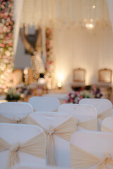 Elegant event seating arrangement with white chairs adorned with light tan ribbons. Flowers and soft lighting create a warm, inviting ambiance.