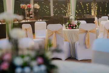 Elegant event setting: white chairs adorned with light gold ribbons surround tables topped with floral centerpieces, set against a backdrop of soft, twinkling lights.