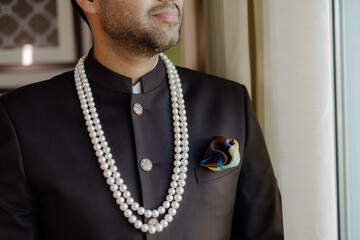 A sharply-dressed man in a dark suit and a pearl necklace is framed in a medium shot. He's ready for a formal event, exudes elegance and confidence.